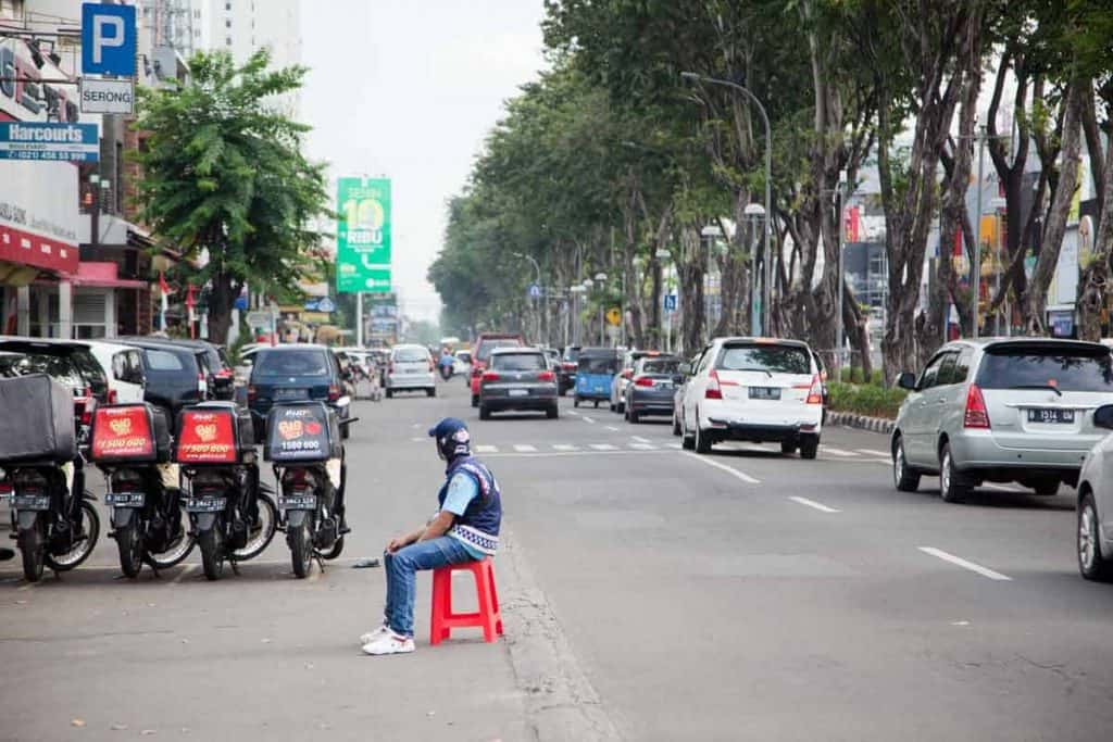 Straße in Jakarta. Ein MAnnn sitzt auf der Fahrbahn mit einem orangen Plastikhocker