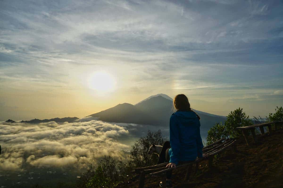 Martina von NomadenStory sitzt bei Sonnenaufgang am Rand des Kraters des Mount Batur und beobachtet die Sonne hinter dem Gunung Abnag und dem Batur Lake aufgehen