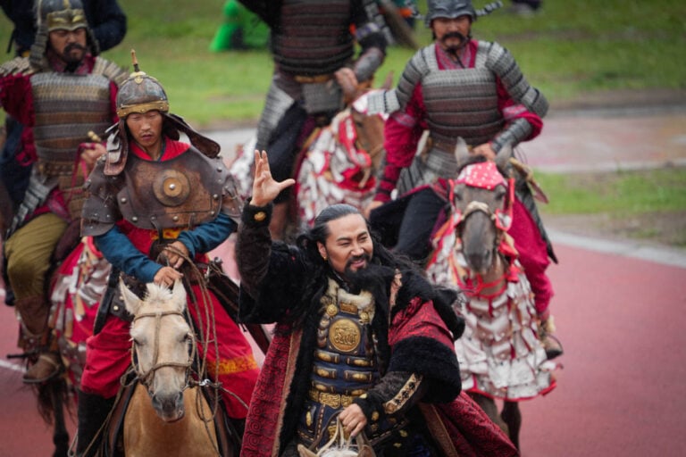 Männer in kunstvollen, traditionellen Kriegerkostümen reiten bei einer Naadam-Parade auf Pferden, einer winkt und lächelt in die Kamera.