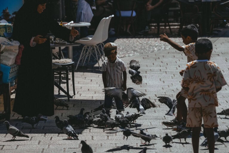 Kinder und eine Frau füttern Tauben auf einer sonnenbeschienenen Straße in Sarajevo. Die Vögel fliegen und picken auf dem Boden.