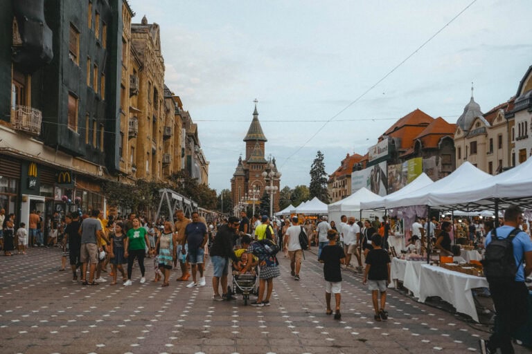 Geschäftiges Treiben auf dem Markt in Timișoara mit Menschen, die zwischen den Ständen umhergehen, umgeben von historischen Gebäuden und einer Kirche im Hintergrund.