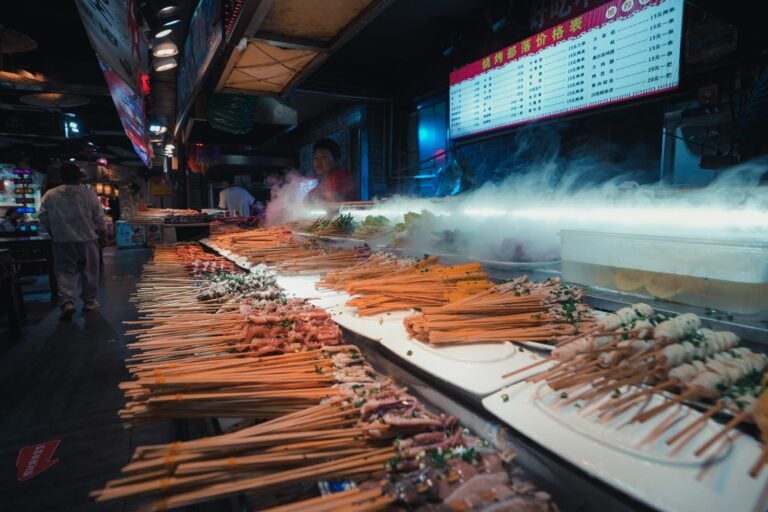 Auf einem belebten Nachtmarkt in China werden Fleisch- und Gemüsespieße angeboten, darüber die Speisekarte.