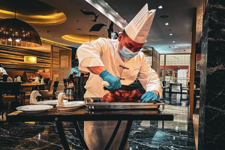 Ein Koch in weißer Uniform und Maske tranchiert Peking-Ente-Braten in einem Restaurant.