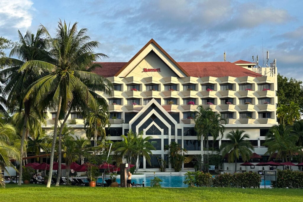 Das Miri Marriott Resort ist ein großes Hotel mit einem markanten roten Dach und mehreren Balkonen, ergänzt durch einen glitzernden Swimmingpool, der von Palmen umgeben ist. Der Himmel ist teilweise bewölkt und wirft sanfte Schatten auf den Rasen im Vordergrund.