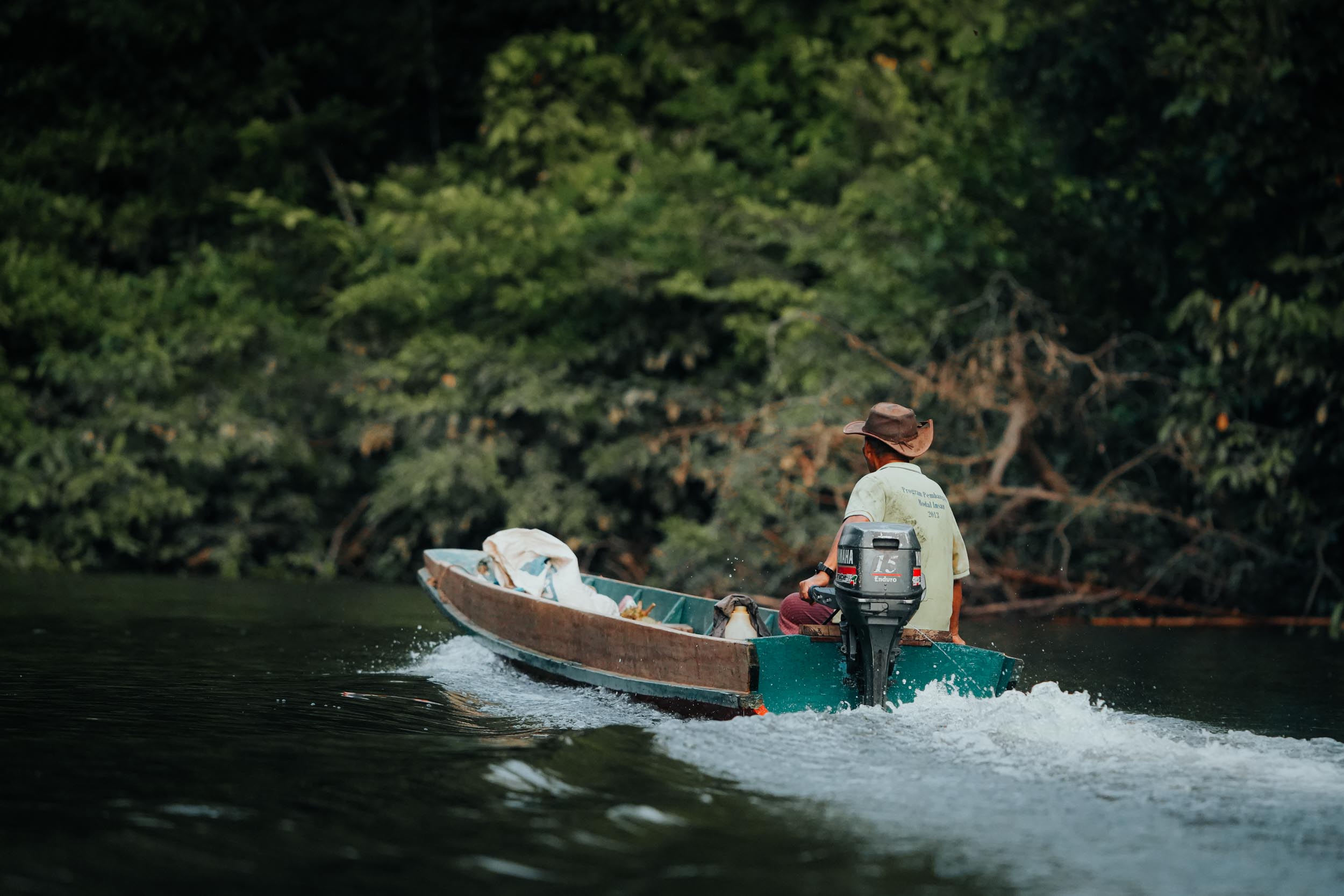 Eine Person mit Hut steuert ein kleines Motorboot entlang eines bewaldeten Flusses.