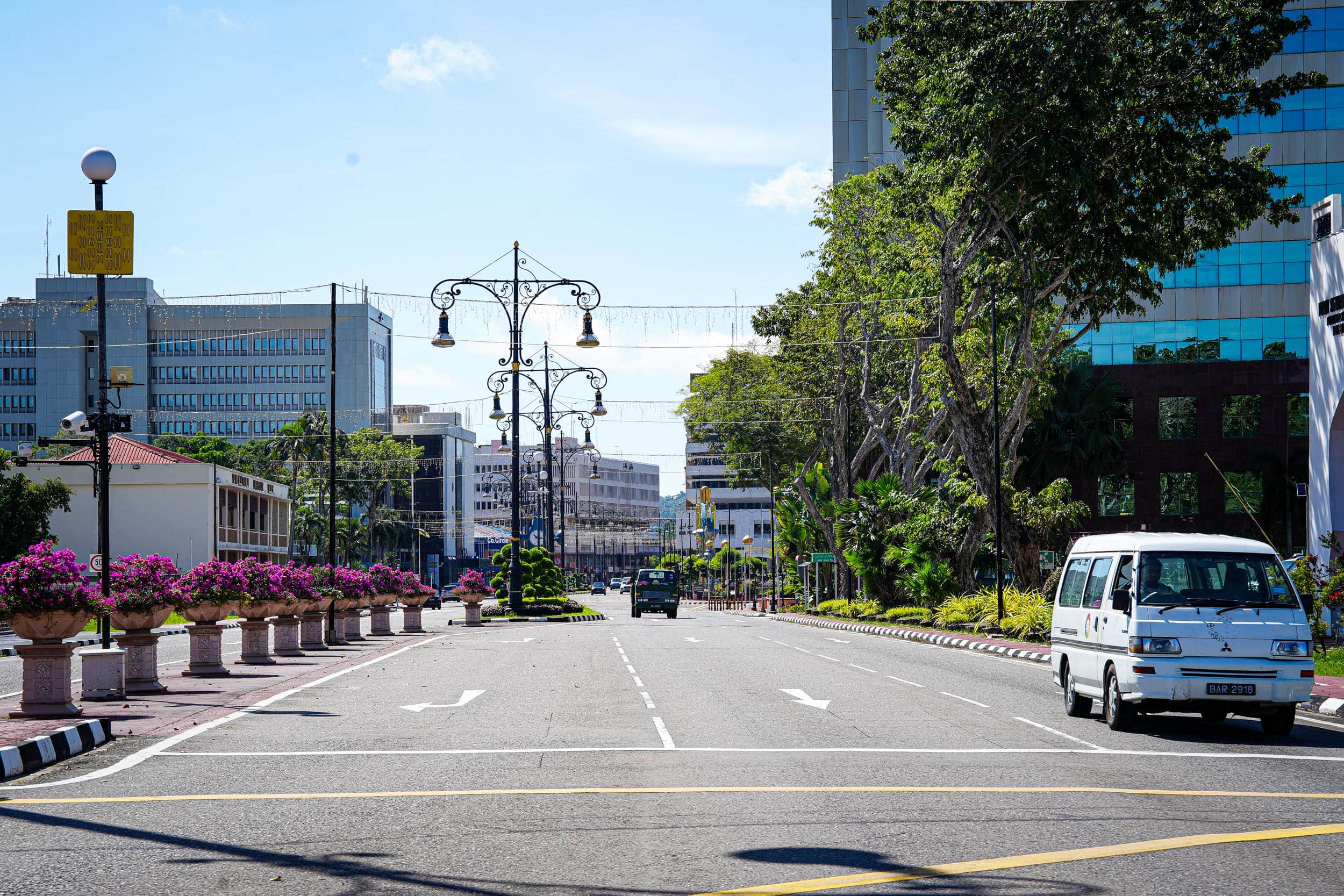 Breite Stadtstraße mit Gebäuden, Bäumen, Straßenlaternen und einem unter einem klaren blauen Himmel auf der Straße geparkten Lieferwagen.