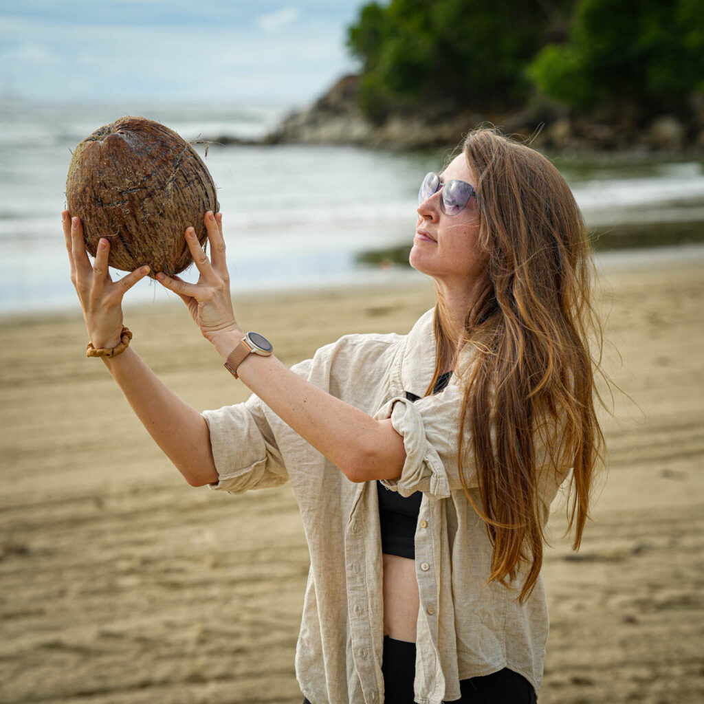 Frau an einem Strand, die eine große Kokosnuss hält, eine Sonnenbrille und ein beiges Hemd trägt, im Hintergrund das Meer.