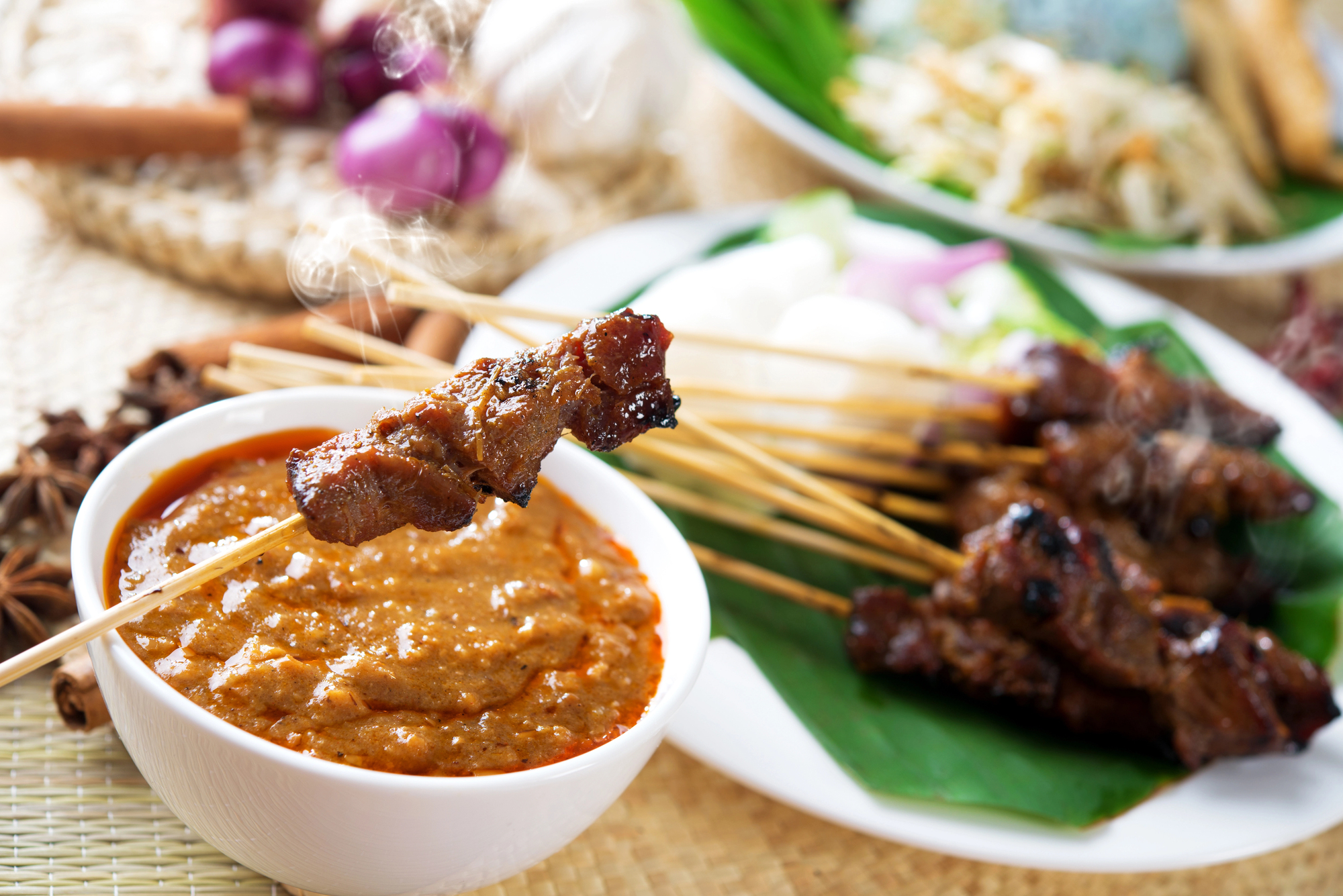 Saté-Spieße mit Erdnusssauce in einer weißen Schüssel, umgeben von frischem Gemüse auf einer geflochtenen Matte.