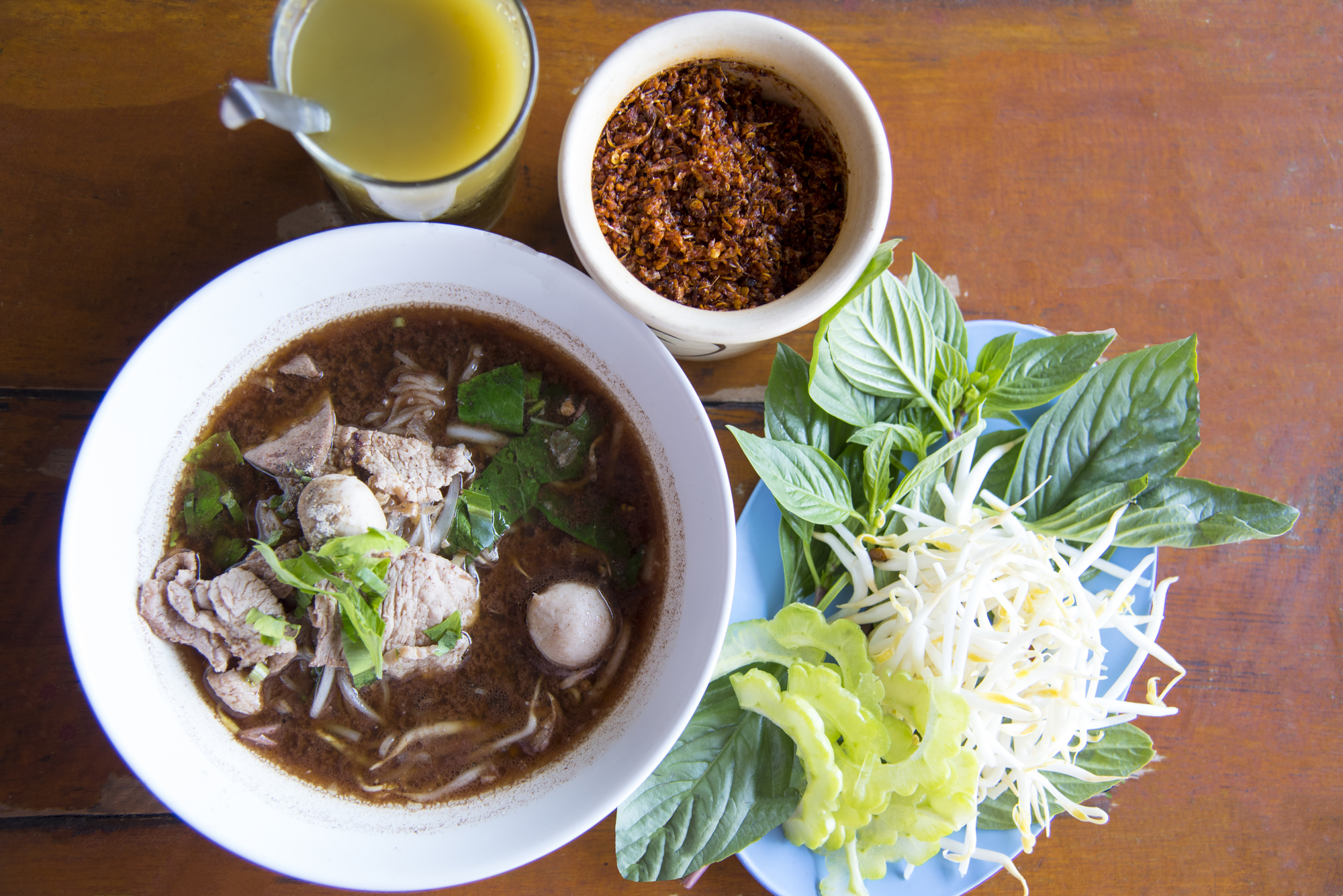 Thailändische Nudelsuppe mit Rindfleisch und Gemüse, dazu frische Kräuter, Sojasprossen, Chiliflocken und ein Glas Saft.