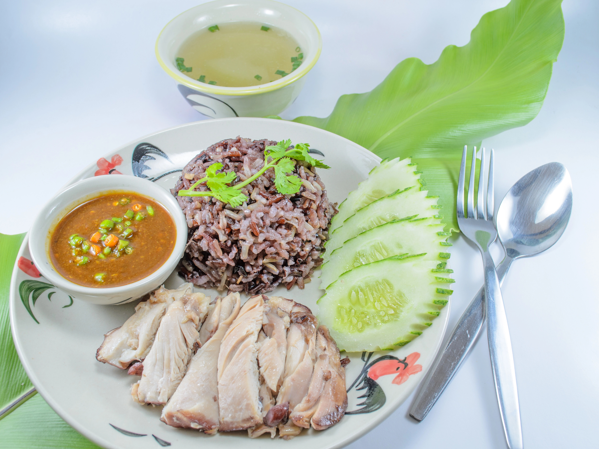 Teller mit Hühnchenscheiben, Reis, Gurke und Soße; Schüssel Suppe; Löffel und Gabel auf Bananenblatt-Hintergrund.