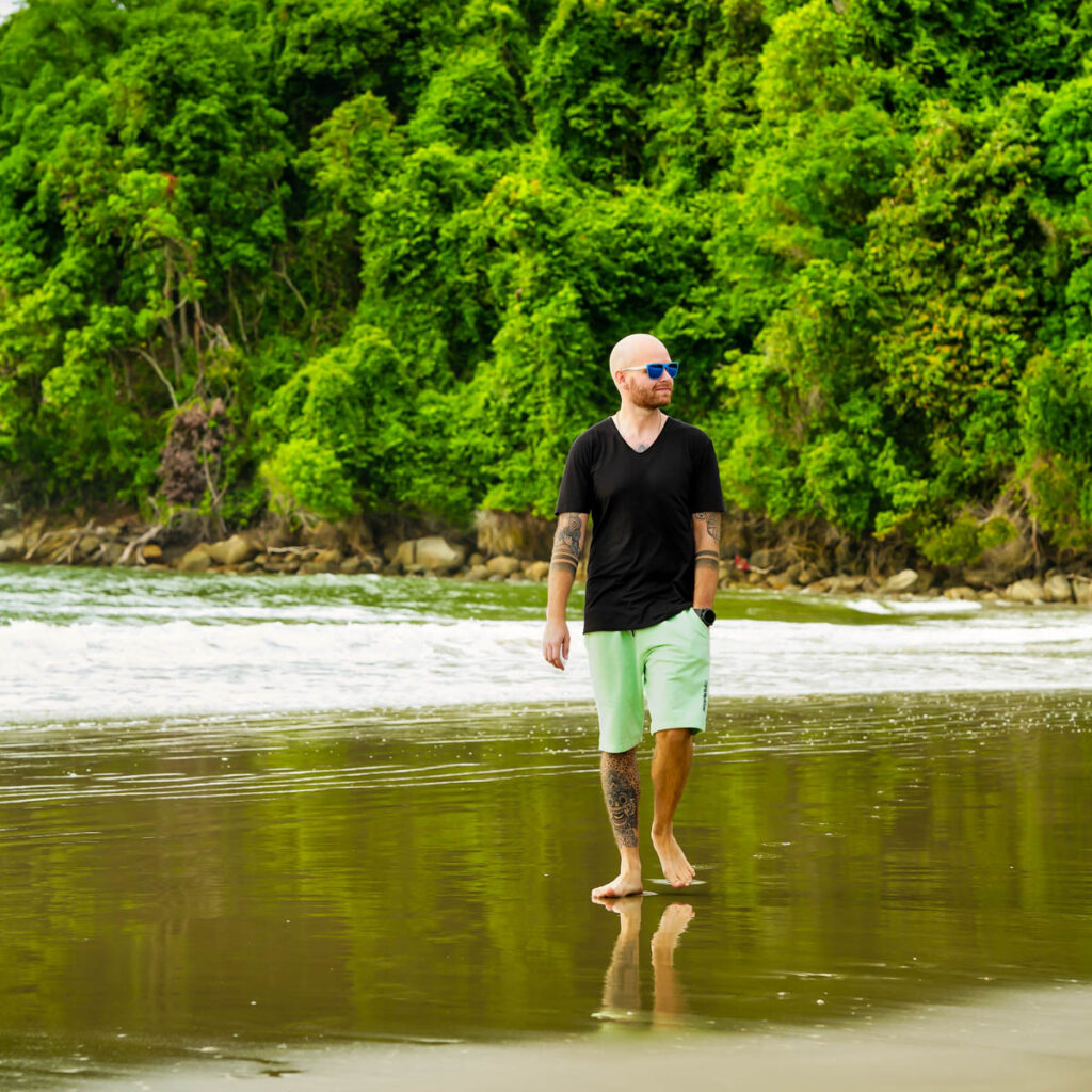 Mann mit Sonnenbrille läuft barfuß an einem Strand entlang. Er trägt ein schwarzes T-Shirt und grüne Shorts, im Hintergrund ist üppiges Grün.