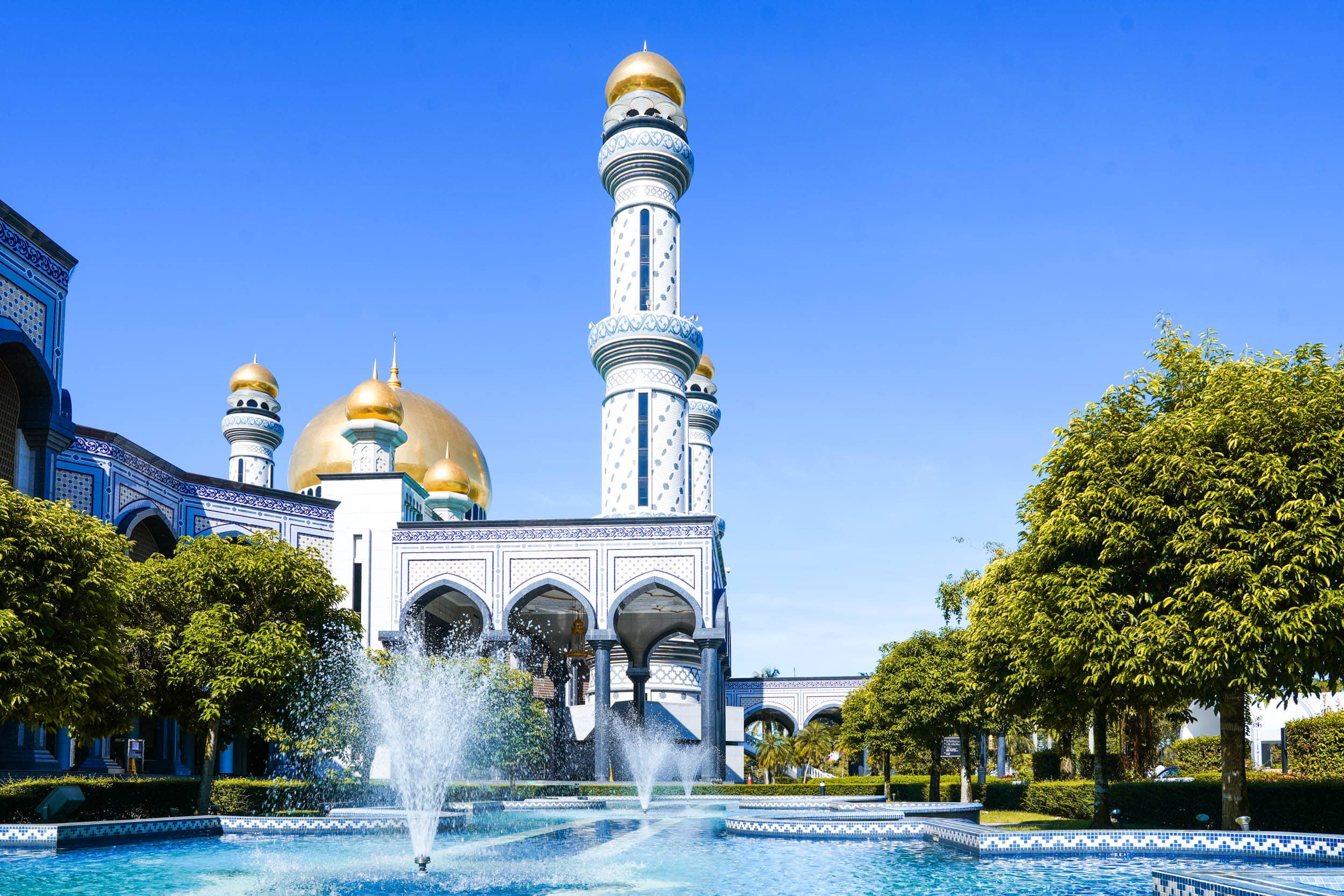 Majestätische Moschee mit goldenen Kuppeln und Minaretten, umgeben von Bäumen und Brunnen unter einem klaren blauen Himmel.