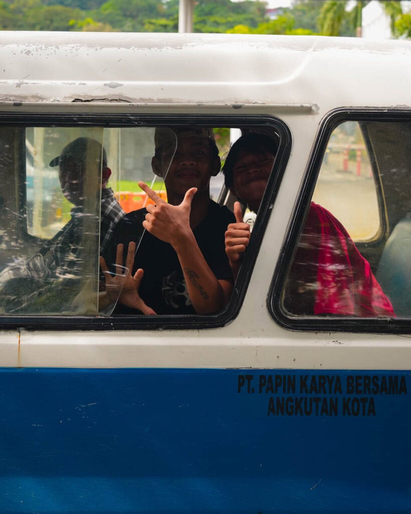 Menschen in einem blau-weißen öffentlichen Nahverkehrswagen in Balikpapan lächeln und gestikulieren mit den Händen durch ein offenes Fenster.