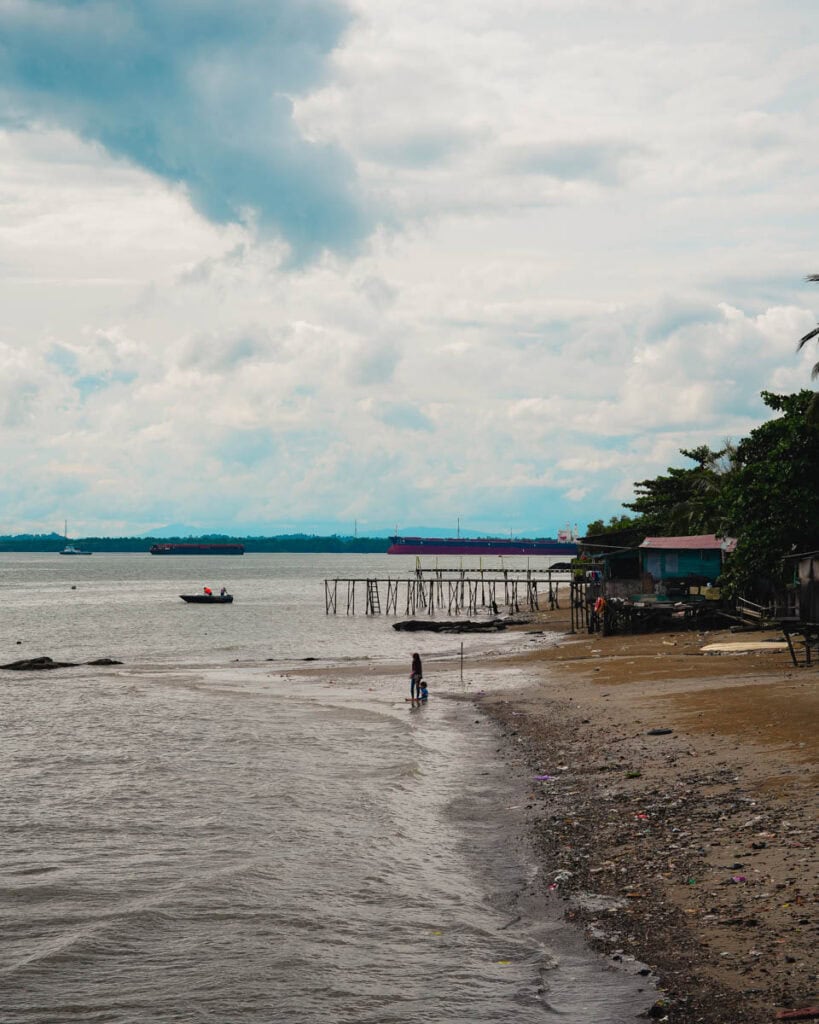 Eine Person mit einem Fahrrad schlendert an einem wolkigen Strand in Balikpapan entlang, wo sich ein Holzsteg erstreckt und in der Ferne ein Boot den Horizont ziert.