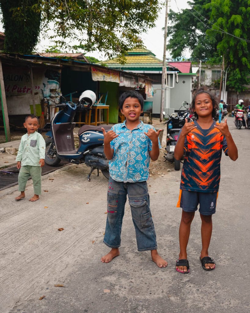 Zwei lächelnde Kinder, die Shaka-Zeichen machen, stehen barfuß auf einer Straße in Balikpapan. Im Hintergrund sind ein weiteres Kind und Motorräder zu sehen.