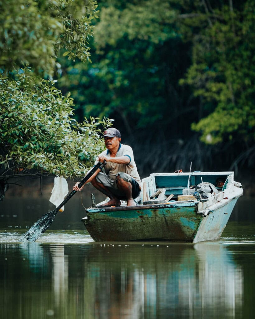 In der üppigen, grünen Flusslandschaft von Balikpapan paddelt ein Mann mit seinem kleinen Holzboot, umgeben von einer ruhigen Baumlandschaft.