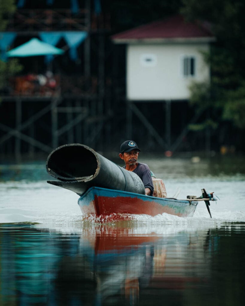 Ein Mann auf einem Boot navigiert durch die ruhigen Gewässer von Balikpapan und transportiert geschickt ein großes Rohr. Im Hintergrund erheben sich anmutig Gebäude auf Stelzen aus dem Wasser und spiegeln die einzigartige Harmonie der Region zwischen Natur und menschlichem Einfallsreichtum wider.