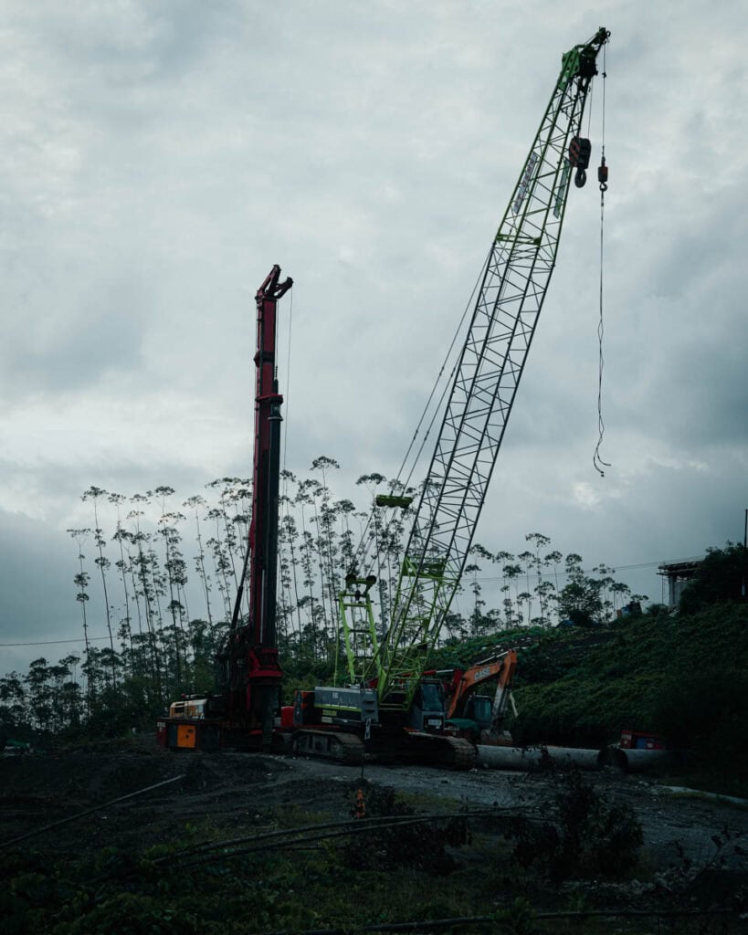 Die Nusantara-Baustelle mit großen Kränen und Maschinen, eingebettet zwischen hohen Bäumen unter einem bewölkten Himmel, zeigt die harmonische Verbindung von Natur und Fortschritt.