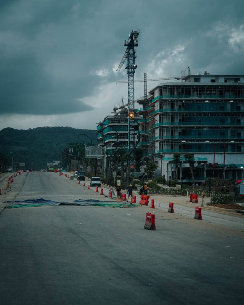 In einer verlassenen Straße in Nusantara sind Hochhäuser und ein Kran inmitten einer Baustelle zu sehen. Unter einem bewölkten Himmel säumen Verkehrsleitkegel den Weg.