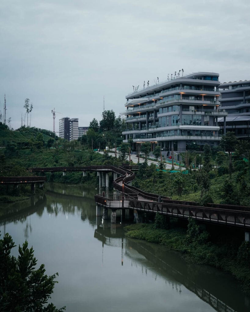 Das moderne Gebäude von Nusantara steht anmutig neben einem gewundenen Fluss und einer Fußgängerbrücke, eingebettet in üppiges Grün unter einem wolkigen Himmel.