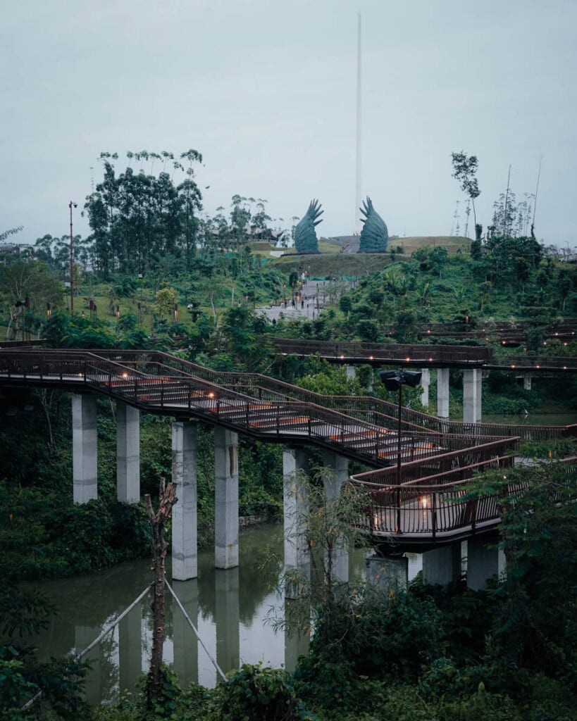 Erhöhter Holzsteg im üppigen Nusantara-Park mit großen Vogelskulpturen und einem hohen Turm im Hintergrund.