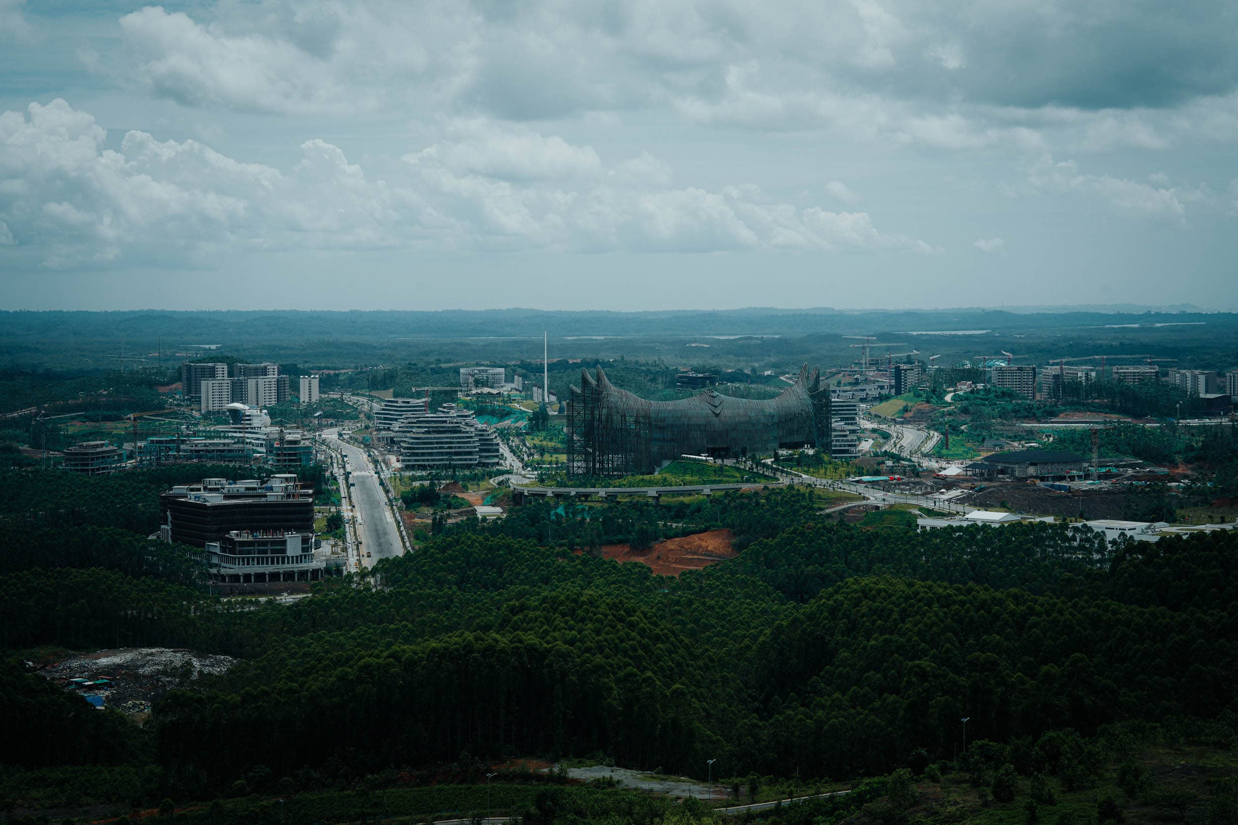 Luftaufnahme der weitläufigen Landschaft von Nusantara mit modernen Gebäuden, verschlungenen Straßen und üppigem Grün unter einem bewölkten Himmel.