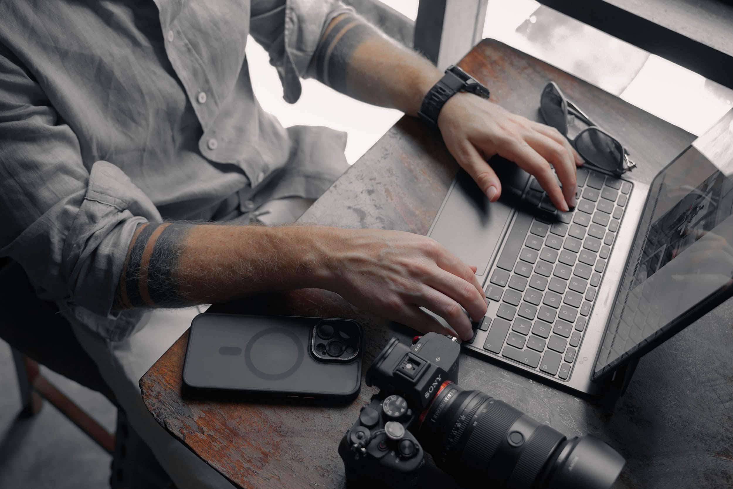 Person, die an einem Holztisch auf einem Laptop tippt, in der Nähe eine Kamera, ein Smartphone und eine Sonnenbrille.