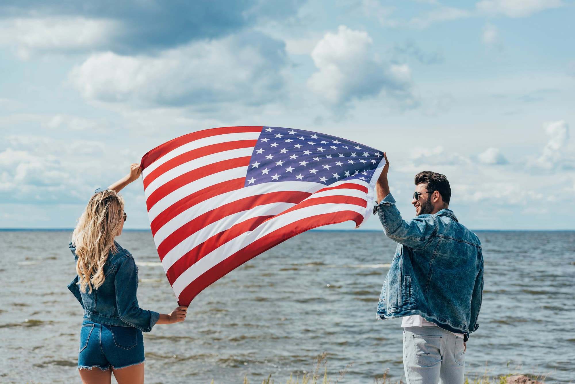 Zwei Menschen halten eine große amerikanische Flagge am Meer, unter einem bewölkten Himmel. Beide tragen Jeansjacken und Sonnenbrillen.