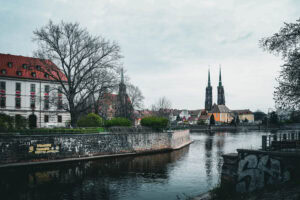 Fluss mit von Bäumen gesäumten Ufern, historischen Gebäuden und einer Kathedrale mit zwei Türmen unter einem bewölkten Himmel.