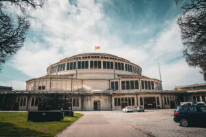 Großes, rundes historisches Gebäude mit vielen Fenstern, einer Flagge darauf und davor geparkten Autos unter einem bewölkten Himmel.
