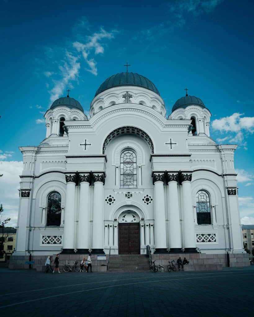 Weiße Kuppelkirche mit Bogenfenstern und Säulen unter blauem Himmel, Spaziergänger und Fahrräder in der Nähe.