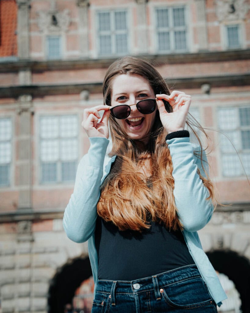 Lächelnde Frau mit Sonnenbrille vor einem historischen Gebäude an einem sonnigen Tag.