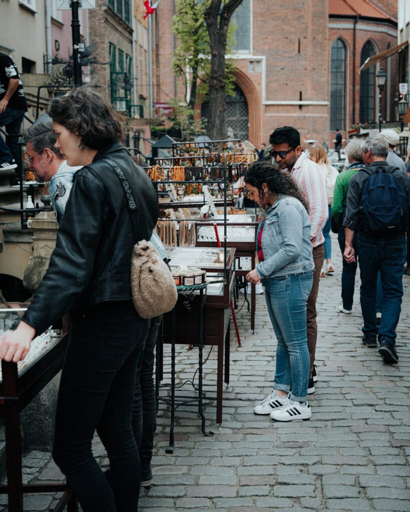 Menschen stöbern an Schmuckständen auf einem Markt im Freien in einer Kopfsteinpflasterstraße in einem historischen Stadtviertel.