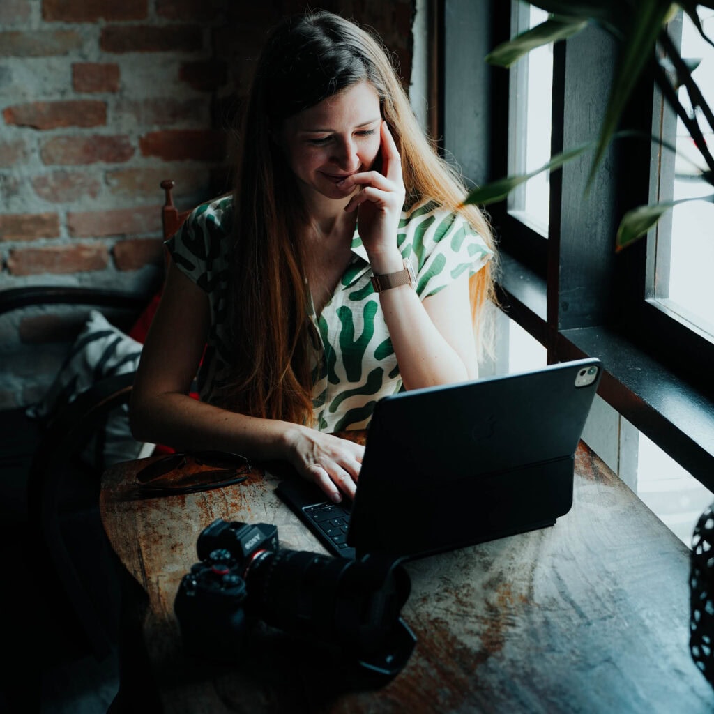Lächelnde Frau, die an einem Holztisch am Fenster sitzt, mit einem Laptop ihr nächstes Abenteuer plant und ihren Weltreise-Blog aktualisiert, mit einer Kamera vor sich.