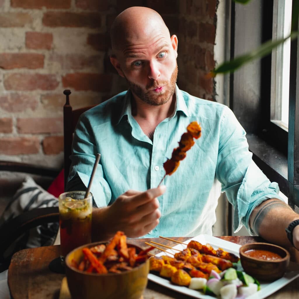 Ein Mann in einem blaugrünen Hemd schneidet ein lustiges Gesicht, während er an einem Restauranttisch einen Spieß mit Essen hält und einen heiteren Moment für seinen Weltreise-Blog festhält.