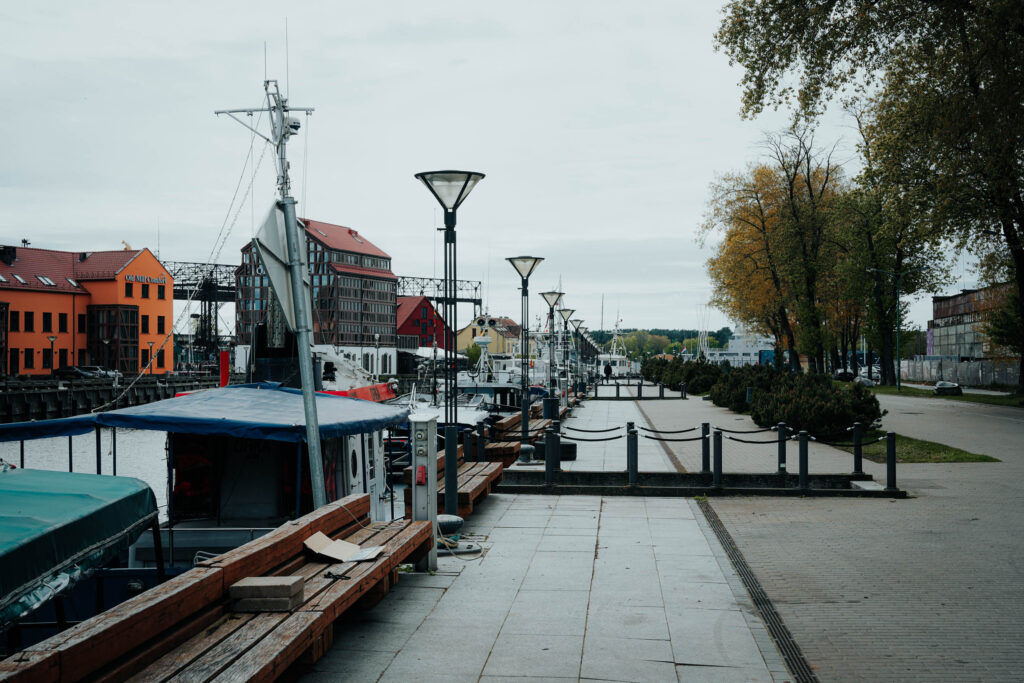 An einem bewölkten Tag legen Boote an einem Uferweg mit Bänken und Bäumen an.