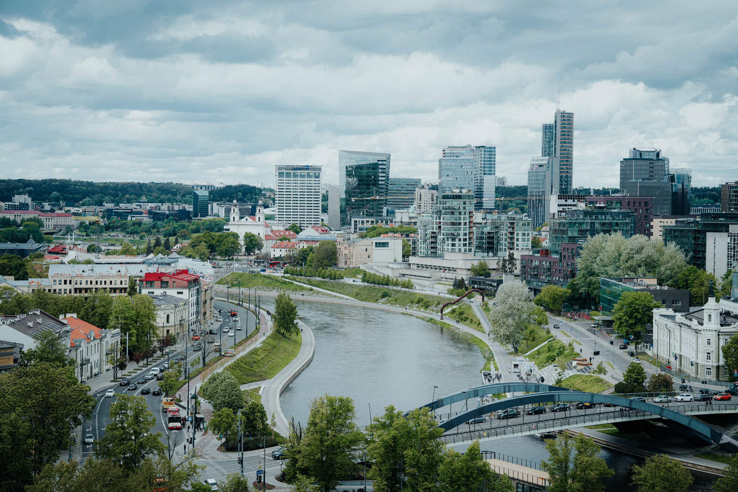 Eine Stadtlandschaft mit modernen Gebäuden, einem Fluss und einer geschwungenen Brücke unter einem bewölkten Himmel.