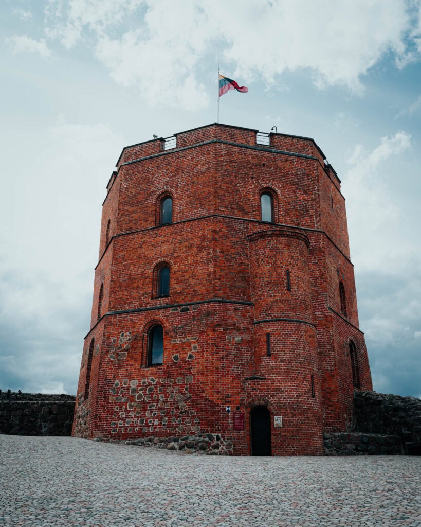 Ein roter Backsteinturm mit einer Flagge auf der Spitze, vor einem bewölkten Himmel und einem gepflasterten Boden.