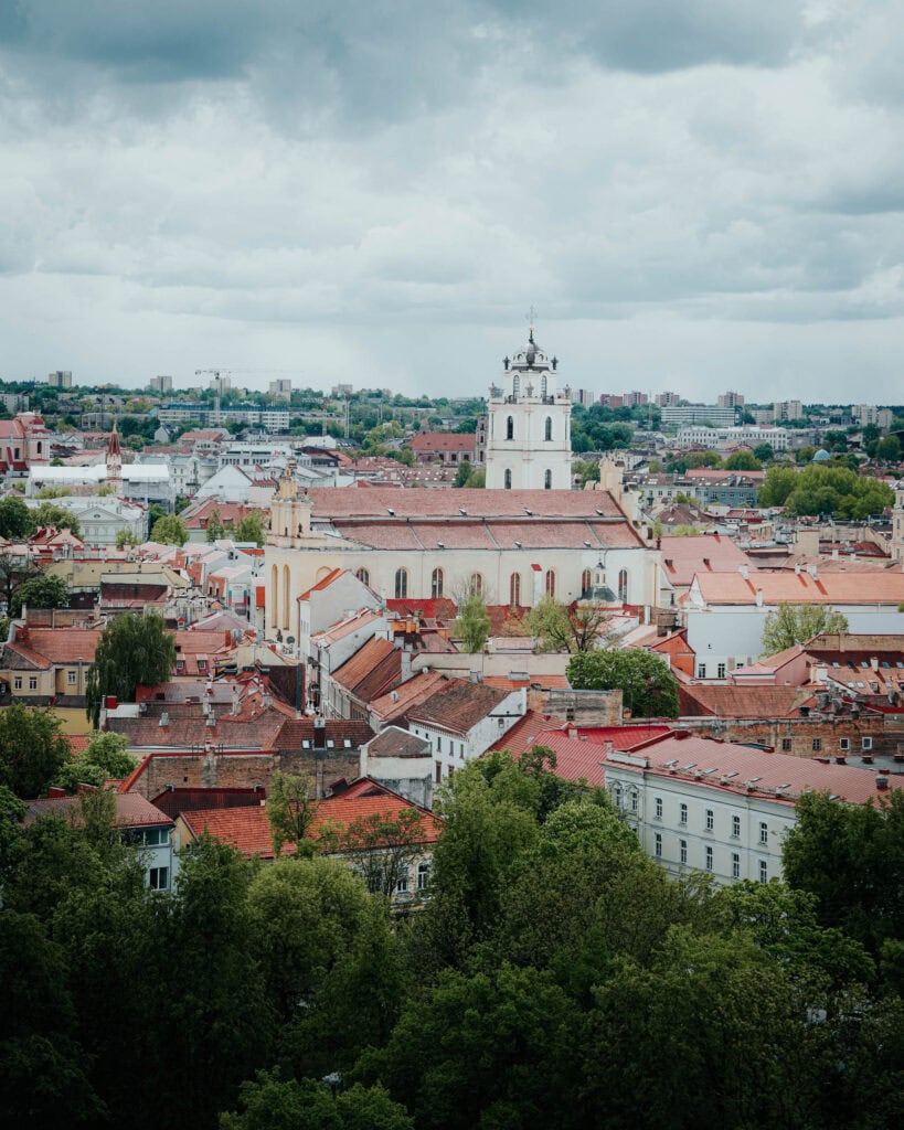 Luftaufnahme einer europäischen Stadt mit roten Dächern, einem Kirchturm und grünen Bäumen unter einem bewölkten Himmel.