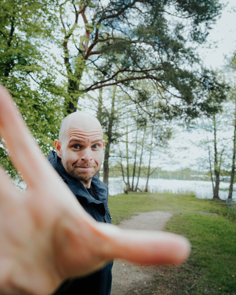 Ein Mann im Freien streckt seine Hand in Richtung Kamera aus, im Hintergrund sind Bäume und ein See zu sehen.