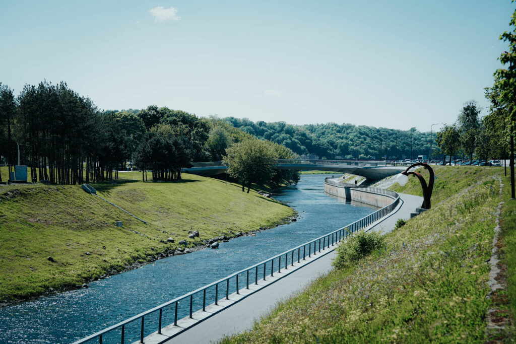 Ein Uferweg mit Geländer, Bäumen und einer Brücke unter einem klaren blauen Himmel.
