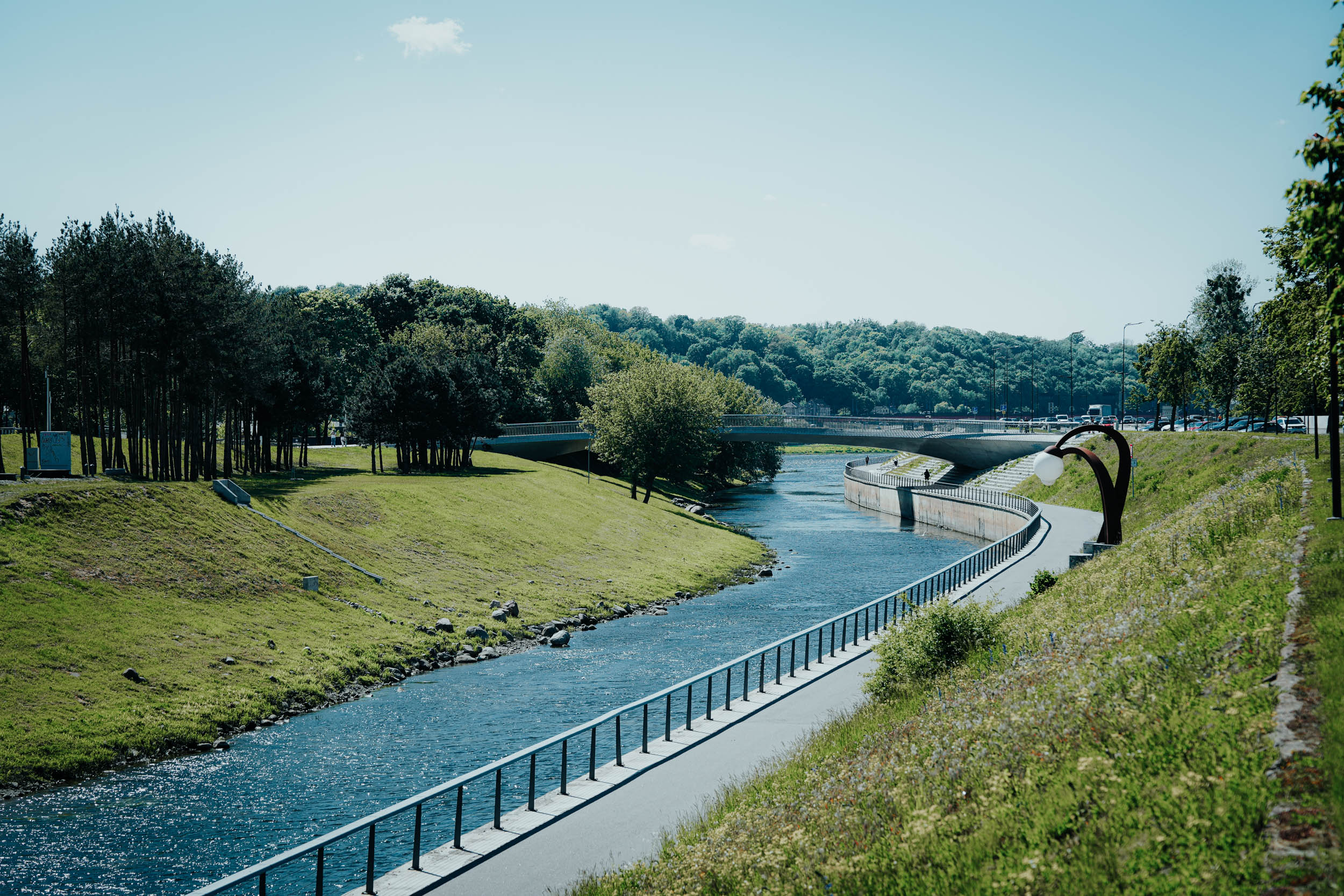 Ein Uferweg mit Geländer, Bäumen und einer Brücke unter einem klaren blauen Himmel.