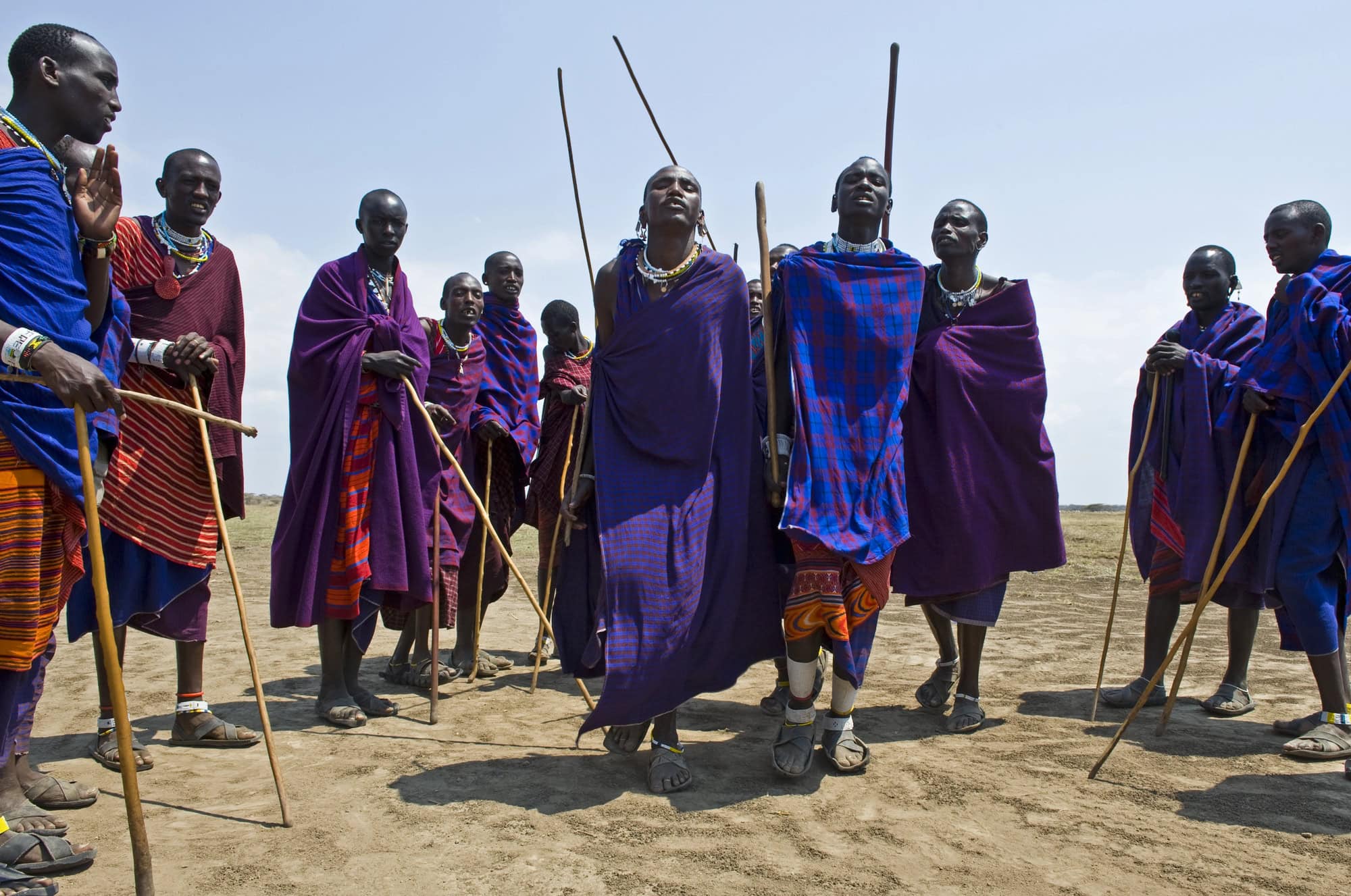Eine Gruppe von Massai in farbenfroher traditioneller Kleidung steht im Freien und hält Holzstäbe.