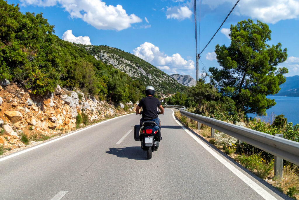 Person, die auf einem Motorroller auf einer kurvenreichen Küstenstraße mit Bergen und blauem Himmel im Hintergrund fährt.