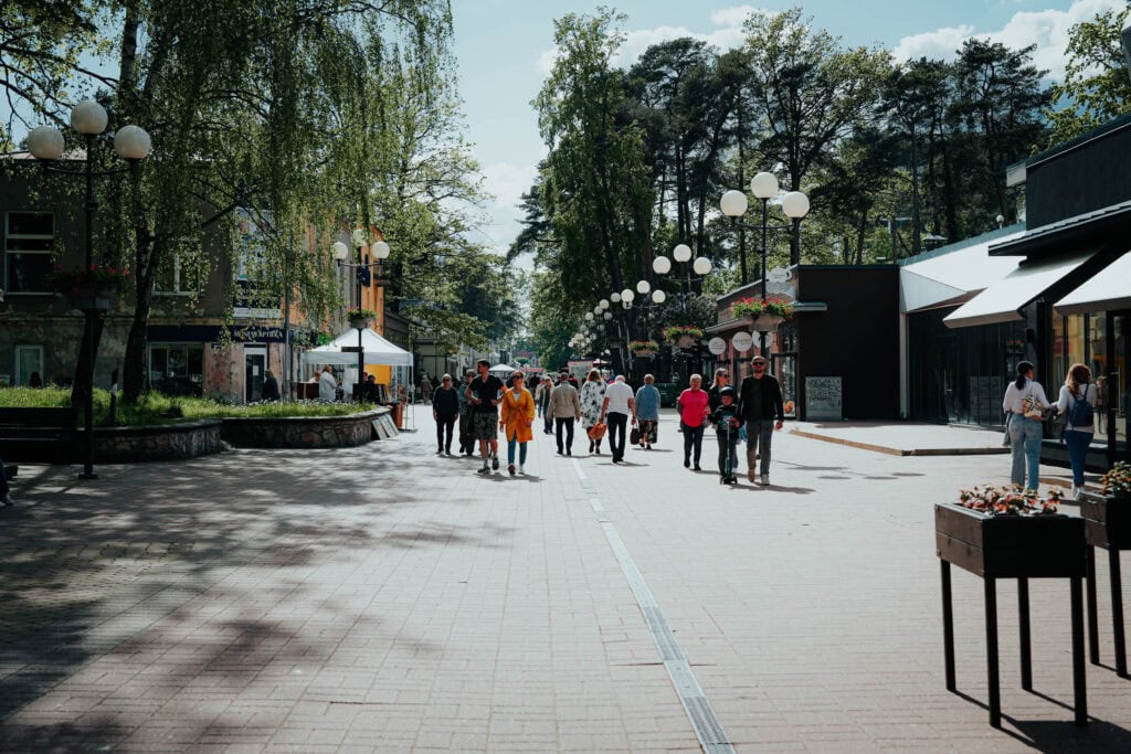Die Menschen gehen eine sonnige, von Bäumen gesäumte Fußgängerzone entlang, die auf beiden Seiten von Geschäften und Außenlaternen gesäumt ist.
