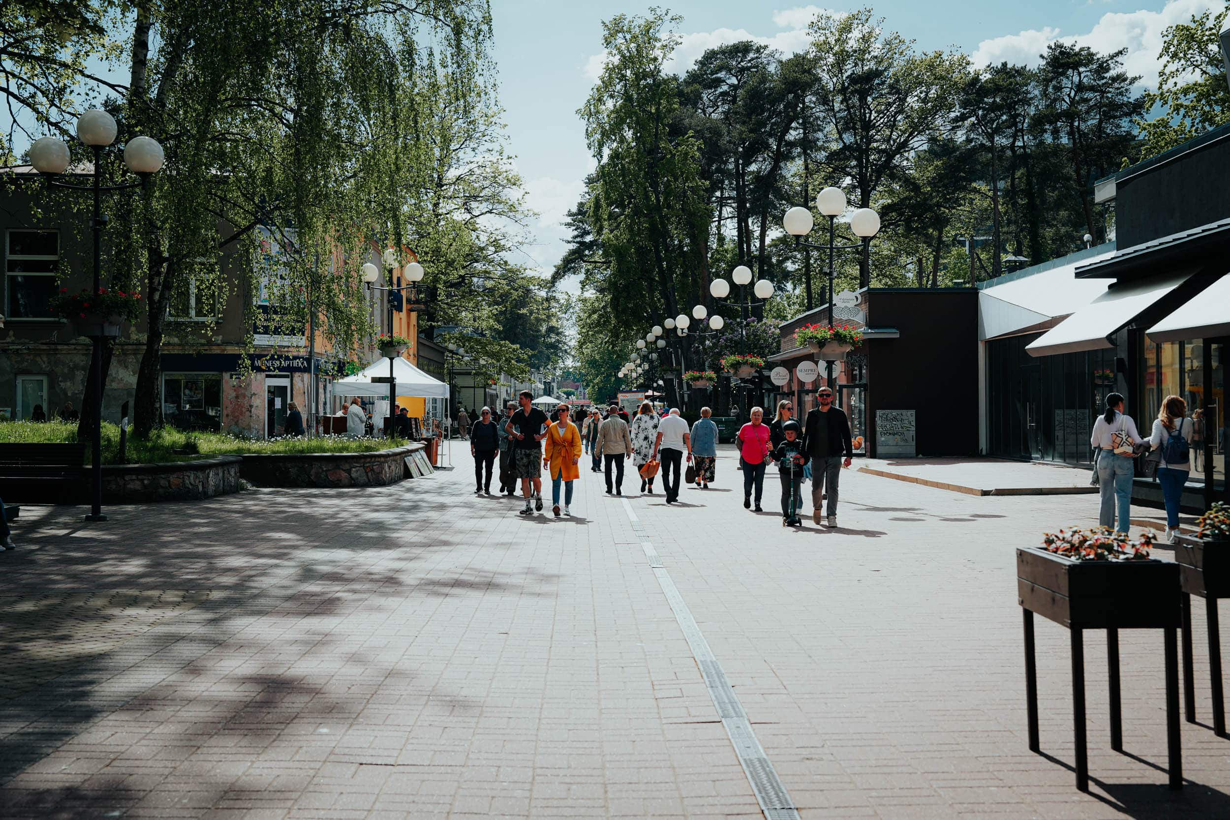 Die Menschen gehen eine sonnige, von Bäumen gesäumte Fußgängerzone entlang, die auf beiden Seiten von Geschäften und Außenlaternen gesäumt ist.