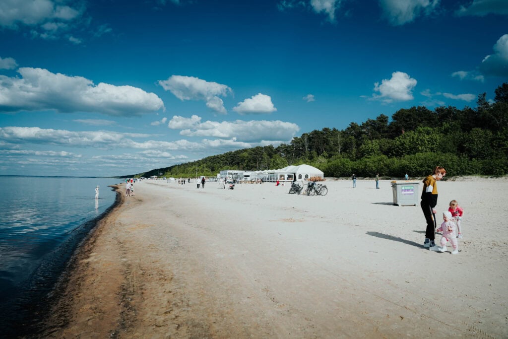 Menschen gehen an einem Sandstrand entlang, im Hintergrund ein Wald, Zelte und ein blauer Himmel mit Wolken.