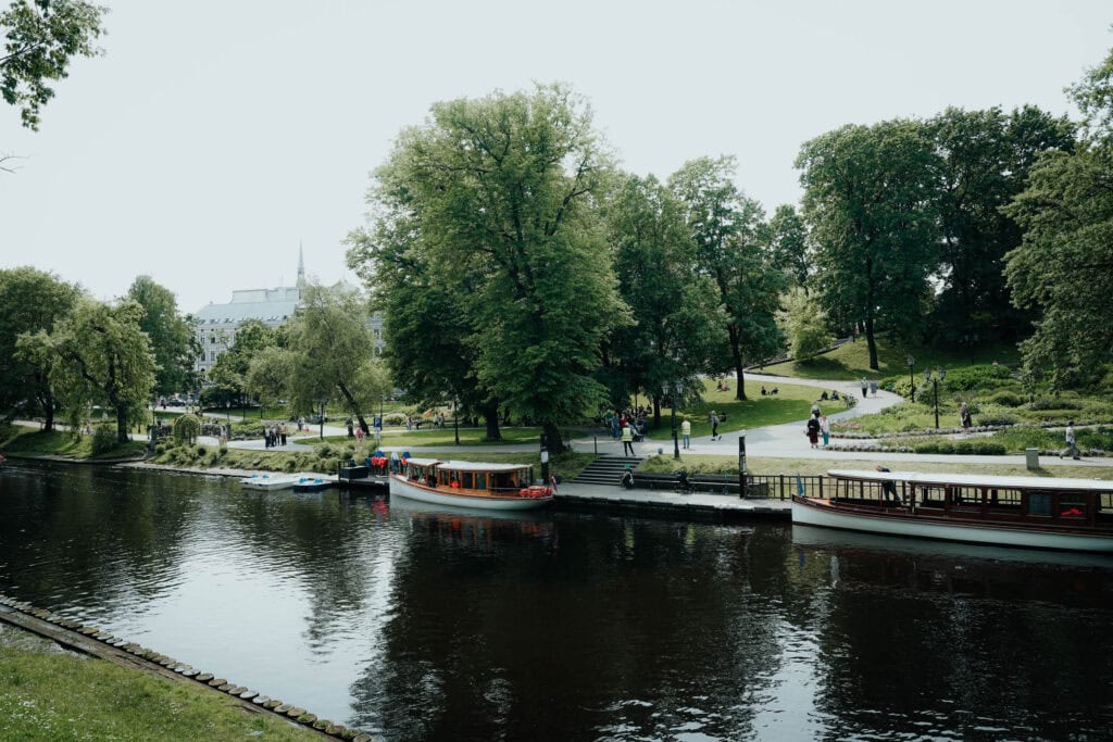 Menschen gehen in einem üppig grünen Park neben einem ruhigen Fluss spazieren und entspannen sich, am Ufer liegen Boote.