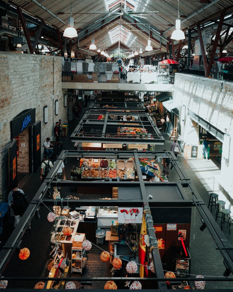 Markthalle in Tallinn mit Essens- und Obstständen, hohen Decken, Käufern und einfallendem Tageslicht.