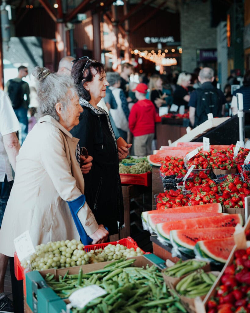 Zwei Frauen stehen an einem Marktstand in Tallinn, umgeben von Erdbeeren, Wassermelonen, Weintrauben und anderen frischen Produkten.
